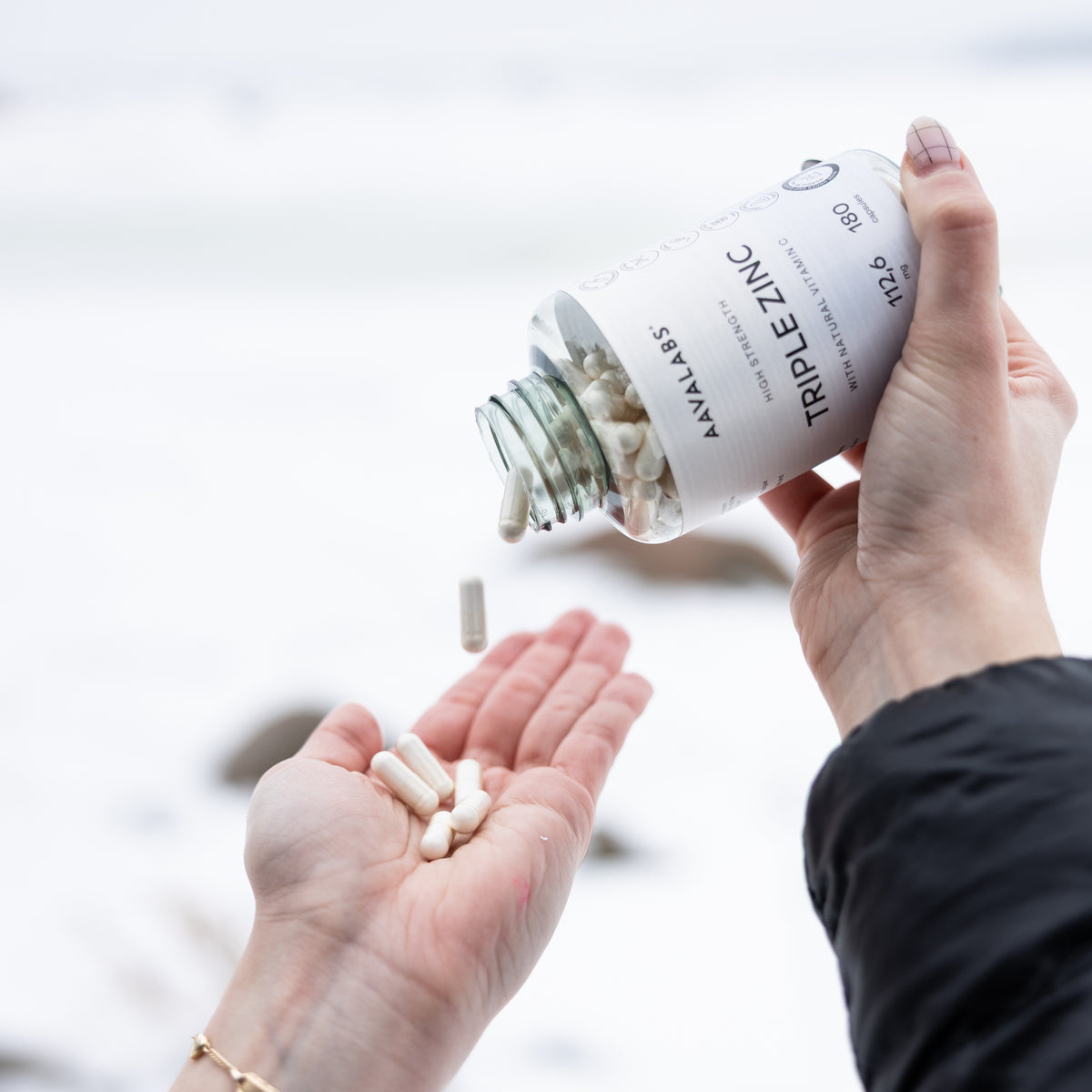 White capsules pouring from a clear glass Aavalabs TRIPLE ZINC bottle into a hand against a snowy background.