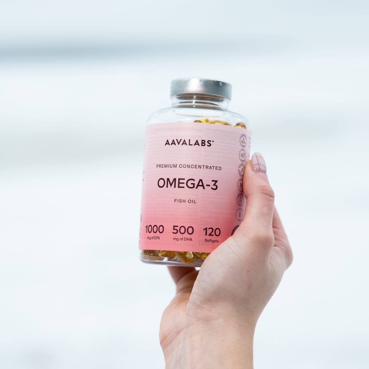 Hand holding a clear plastic bottle of Aavalabs OMEGA-3 fish oil softgels with a pink label against a blurred background.